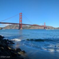 海岸沿いから見たゴールデンゲートブリッジ。青空の下、穏やかな波が手前の砂浜に打ち寄せ、左手には黒い岩場が続く。赤い橋のシルエットが湾を横切り、背景には茶色い丘が広がっている。