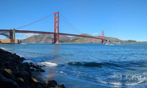 海岸沿いから見たゴールデンゲートブリッジ。青空の下、穏やかな波が手前の砂浜に打ち寄せ、左手には黒い岩場が続く。赤い橋のシルエットが湾を横切り、背景には茶色い丘が広がっている。