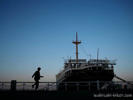 夕暮れの山下公園で係留中の氷川丸を背景に、遊歩道を歩く人物のシルエットを写したセルフスナップ。