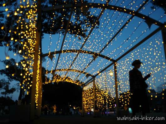 横浜 山下公園のイルミネーションによる光のトンネルの下で立つ人物のシルエット。ブルーアワーの空と無数の電飾が広がるセルフスナップ。