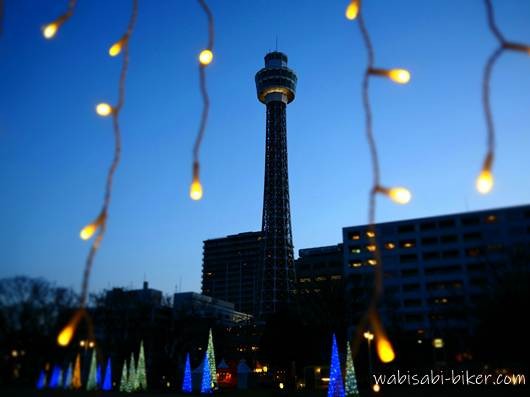 夕暮れの横浜マリンタワーを中心に、足元に並ぶ光のオブジェと周囲の建物を写した風景。手前にはイルミネーションの電球が写り込んでいる。