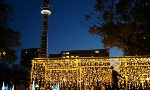 横浜マリンタワーを背景に、イルミネーションの光のトンネルの外側を歩く人物のシルエットを写したセルフスナップ。周囲に光の装飾と建物が見えるブルーアワーの夕景。