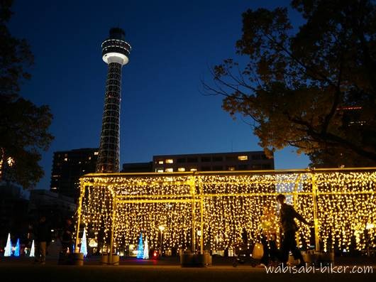 横浜マリンタワーを背景に、イルミネーションの光のトンネルの外側を歩く人物のシルエットを写したセルフスナップ。周囲に光の装飾と建物が見えるブルーアワーの夕景。