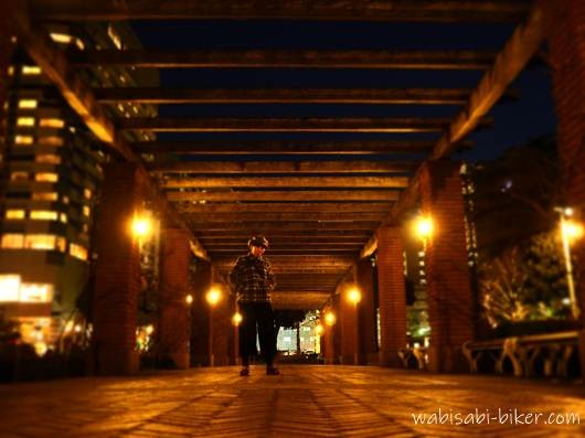 夜の山下公園のパーゴラで、レンガ柱と木製の梁が並ぶ通路の中央に立つ人物のセルフスナップ。両側に街灯と建物の明かりが見える。