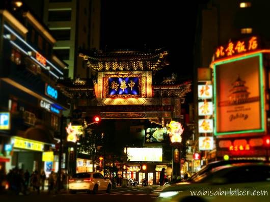 夜の横浜中華街に立つ朝陽門と周囲のネオン看板。道路を走る車と歩行者が写る街景。