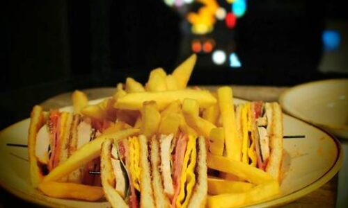 ローズホテル横浜の客室で提供されたルームサービスのクラブハウスサンドとフライドポテトのプレート。背後の窓の外には、ぼかされた中華街の夜景の光が見える。