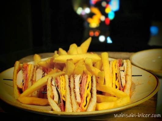 ローズホテル横浜の客室で提供されたルームサービスのクラブハウスサンドとフライドポテトのプレート。背後の窓の外には、ぼかされた中華街の夜景の光が見える。