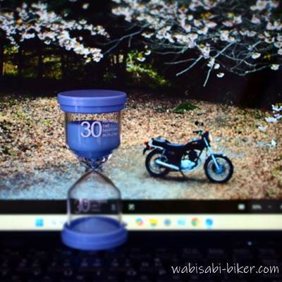 黒いノートパソコンのキーボード手前に青色の30分砂時計が置かれ、画面には桜の前に停めた黒いバイクSR125の壁紙が表示されている。