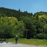 初夏の青空の下、左に黒いバイクSR125を停め、人物がバイクへ向かって歩くセルフスナップ。奥には緑の山が広がり、一部が黄色く色づいている。草地と砂利の広場が手前に続く風景。