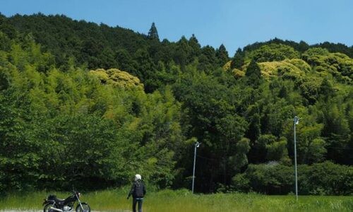 初夏の青空の下、左に黒いバイクSR125を停め、人物がバイクへ向かって歩くセルフスナップ。奥には緑の山が広がり、一部が黄色く色づいている。草地と砂利の広場が手前に続く風景。