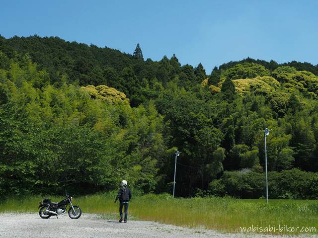 初夏の青空の下、左に黒いバイクSR125を停め、人物がバイクへ向かって歩くセルフスナップ。奥には緑の山が広がり、一部が黄色く色づいている。草地と砂利の広場が手前に続く風景。