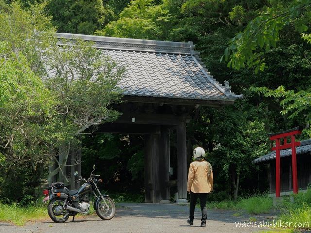 初夏の緑に囲まれた山門の前で、黒いバイクSR125を左に停め、背中を向けて山門を見上げるバイク乗りのセルフスナップ。右奥には赤い鳥居が見える境内の入口。