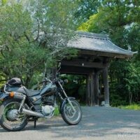 初夏の緑に囲まれた山門の前に、黒いバイクSR125が左に停められている。木陰と日差しが入り混じる静かな境内入口の風景。