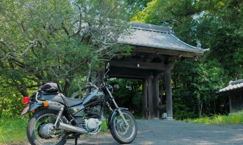初夏の緑に囲まれた山門の前に、黒いバイクSR125が左に停められている。木陰と日差しが入り混じる静かな境内入口の風景。