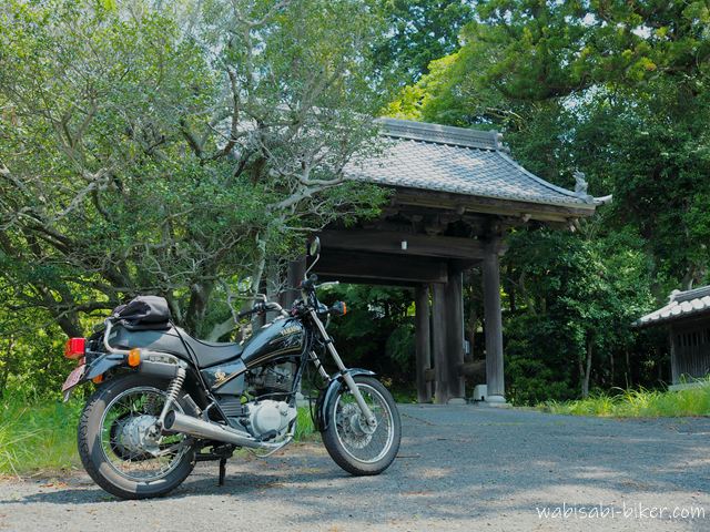 初夏の緑に囲まれた山門の前に、黒いバイクSR125が左に停められている。木陰と日差しが入り混じる静かな境内入口の風景。