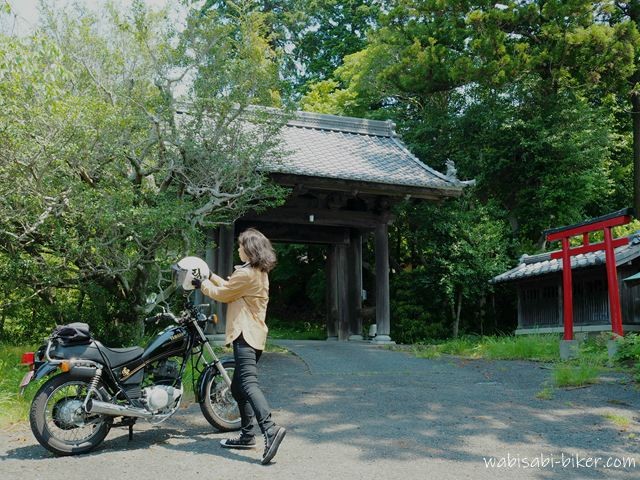 初夏の緑に囲まれた山門の前で、黒いバイクSR125の横に立ち、ヘルメットに手をかけた女性バイク乗りのセルフスナップ。右奥には赤い鳥居が見え、木陰と日差しが混ざる境内入口の風景。
