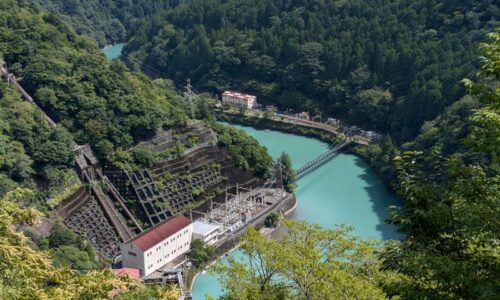 県道から見下ろした奥泉発電所と井川線のアプトいちしろ駅。緑の山々に囲まれた大井川に吊り橋がかかる風景。