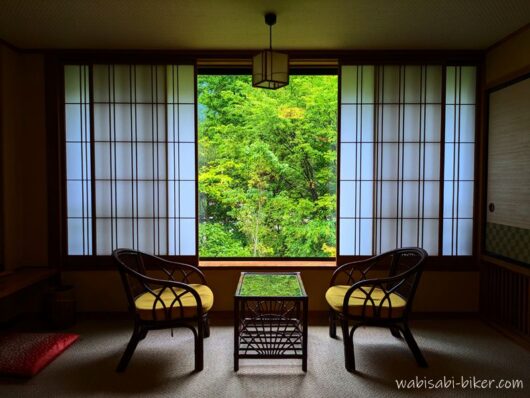 翠紅苑の和室の広縁。窓から少し色づいた緑の木々を望む。窓の緑がテーブルにも少し映り込んでいる。