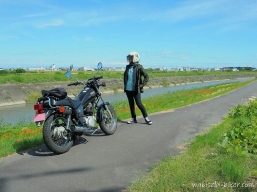ヒガンバナが咲く川沿いで、黒いバイクSR125とヘルメット姿の乗り手を撮ったセルフスナップ。