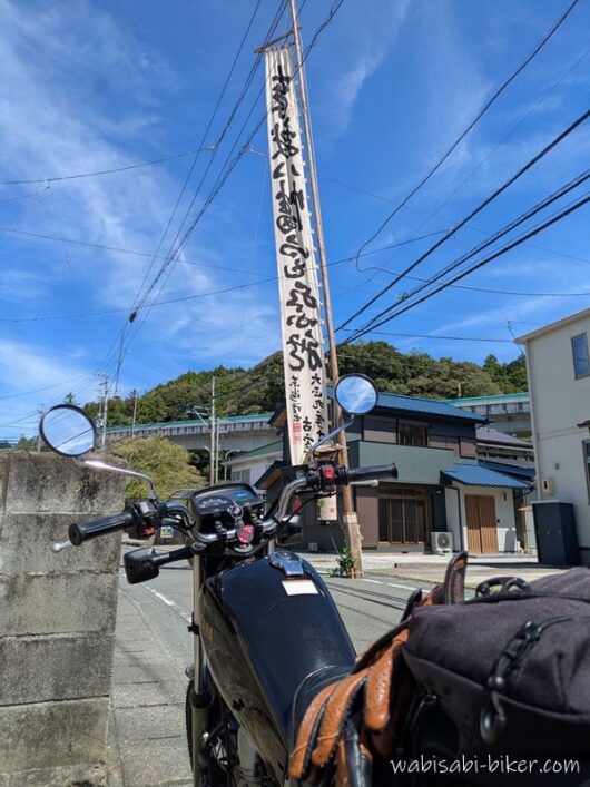 事任八幡宮の祭りの幟が立つ道沿いに停めた、黒いバイクSR125。青空の下、幟旗と住宅地が写っている。