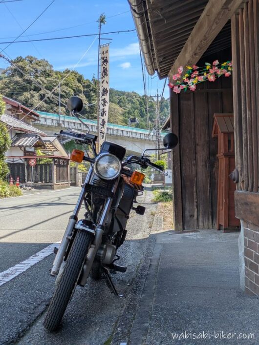 軒花が飾られたレトロ建築「藤文」の前に停めた黒いバイクSR125。奥には祭りの幟旗が立っている。