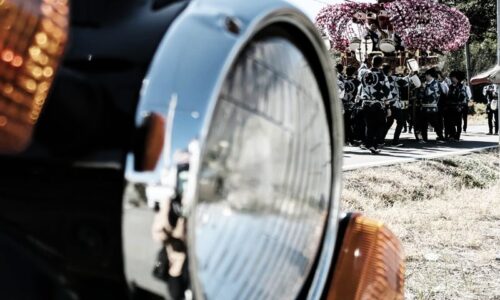 手前に大きく写る黒いバイクSR125の丸いヘッドライトとウインカー。その奥では、ピンク色の花飾りで彩られた祭りの屋台を、大勢の法被姿の人たちが曳いている。