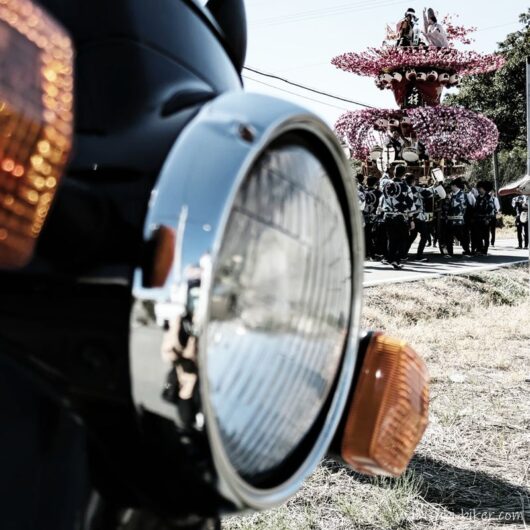 手前に大きく写る黒いバイクSR125の丸いヘッドライトとウインカー。その奥では、ピンク色の花飾りで彩られた祭りの屋台を、大勢の法被姿の人たちが曳いている。
