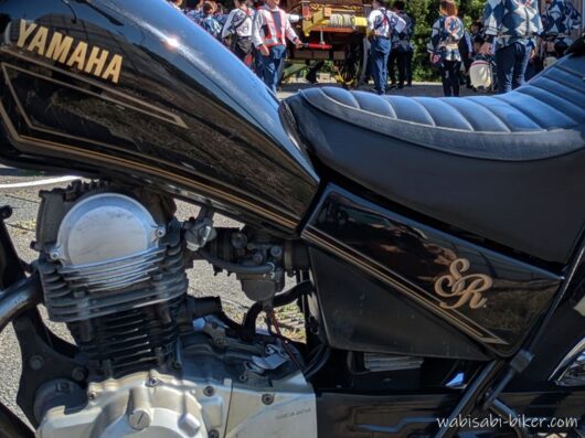 ヤマハSR125の車体側面。燃料タンクの下にエンジンがあり、シート下には金色のSRロゴが入ったサイドカバーが見える。背景には祭りの法被姿の人たち。