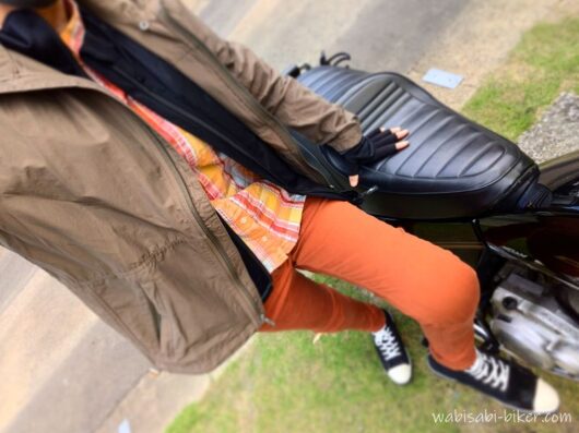 バイクの横に立ち、茶系のウインドブレーカーとオレンジ系のチェック柄シャツ、テラコッタ色のパンツを着用した自撮り。