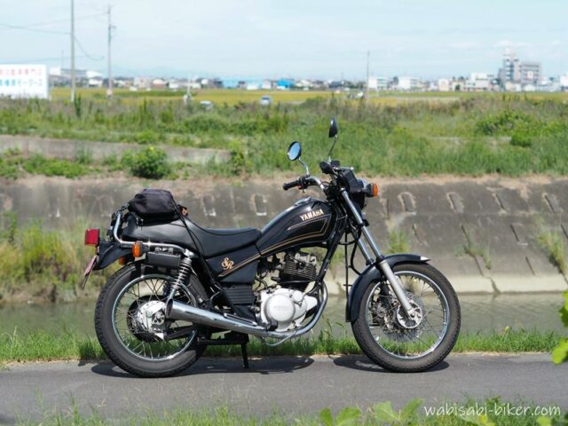 川沿いの道に停めた黒いヤマハSR125。田園風景を背景にしたバイクのサイドビュー。