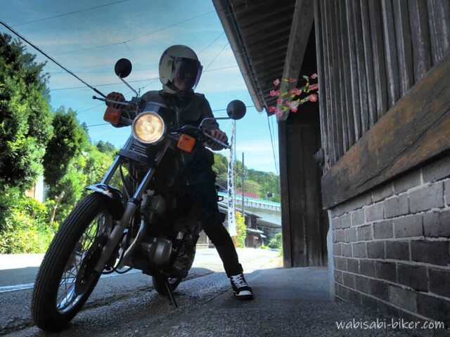 日坂宿の藤文脇に停めた黒いバイクSR125と、ヘルメット姿の乗り手のセルフスナップ。軒先には祭りの花飾りが見える。